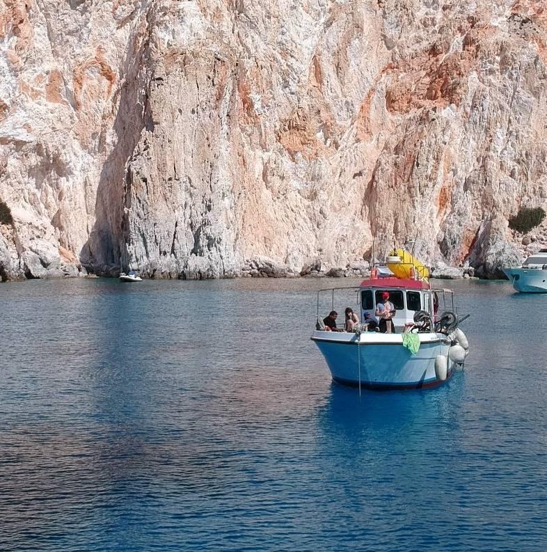A traditional Milian kaïki fishing boat moored beneath white volcanic cliffs, guests aboard